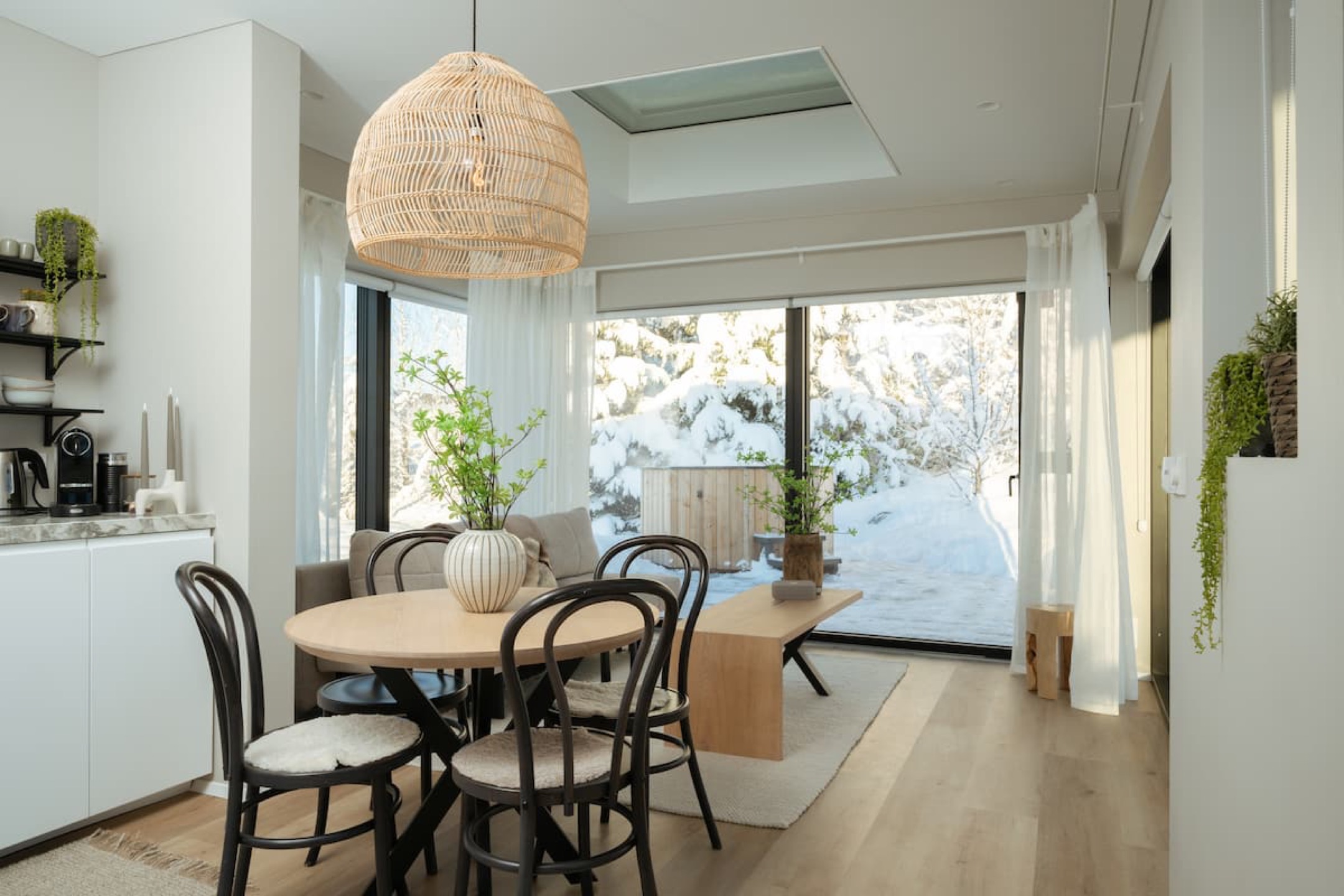 The round dining table under a woven pendant, glass door onto the snow behind it
