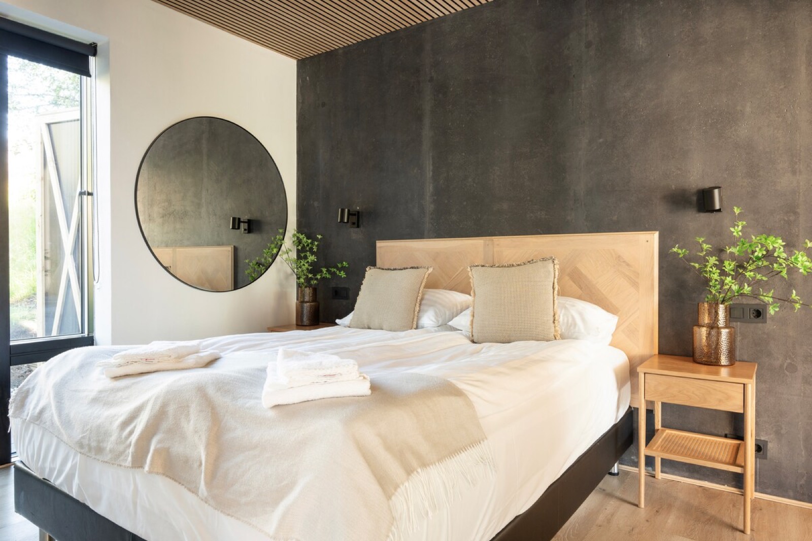 A bedroom with a timber headboard against a dark plaster wall, round mirror beside it